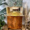 A stylish vintage Rivington Cocktail Cabinet, featuring detailed wood grain patterns and a glass display with a lit interior, stands against a white brick wall adorned with botanical wallpaper. The green and gold Art Deco piece from the 1950s is accentuated by ornate decor and surrounded by plants and pampas grass in vases, enhancing the aesthetic appeal.