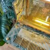 The Rivington Cocktail Cabinet with its tropical monstera leaf print interior and gold-striped glass shelves is open. Warm lighting inside the cabinet accentuates its design. A large green plant with broad leaves is partially visible beside the cabinet on a wooden floor.