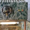 The Rivington Cocktail Cabinet showcases a striking decorative wall with a vintage tropical leaf pattern in silver on a dark green backdrop. A silver ventilation fan is positioned on the left, while four small colored balls are neatly aligned on the right. Beneath it, a lit display shelf with an elegant gold-striped metal surface completes the iconic Art Deco look of this 1950s-style retro bar unit.