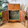 The Rivington Cocktail Cabinet, a vintage walnut drinks bar from the 1950s, is shown with its doors open to reveal an interior adorned with green and white monstera leaf wallpaper. This retro Art Deco unit features elegant brass accents and is complemented by surrounding plants and a fur rug. The setting includes a white brick wall and a decorative sunburst mirror in the backdrop.