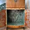 The Rivington Cocktail Cabinet, a vintage walnut drinks unit from the 1950s, stands open to reveal an interior lined with green wallpaper adorned with a leafy Monstera pattern. The outer doors also exhibit a botanical design. Partially visible in the background are indoor plants and a decorative wall mirror, enhancing its retro Art Deco charm.