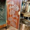 An open door of the Rivington Cocktail Cabinet reveals an intricate design of Monstera leaves. Adjacent to it, a golden decorative lantern featuring geometric cutouts with a white candle is set on the walnut wooden floor. The top of the cabinet is adorned with dry grass decorations, adding a touch of natural elegance.