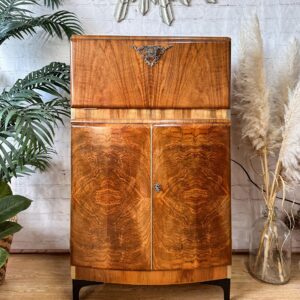 The Rivington Cocktail Cabinet, an Art Deco vintage drinks bar from the 1950s, features a stunning walnut finish with intricate monstera leaf patterns in green and gold. This retro bar unit boasts a decorative metal element at the top and double doors at the bottom. It is elegantly placed against a white brick wall, framed by lush green plants on the left and dried pampas grass in a vase on the right.