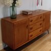 A wooden sideboard with mid-century modern design stands against a white wall. It features two doors and three drawers. On the sideboard are a green vase with white flowers, a decorative box, and two candlesticks with red and beige candles.