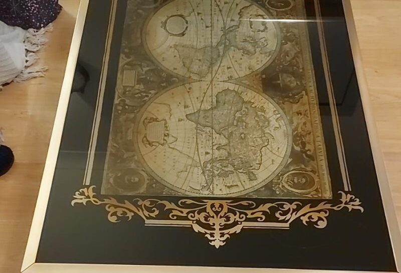 A rectangular wooden coffee table with a glass top featuring an antique-style world map. The map is decorated with ornate, golden designs and borders, and it is centered on the table's surface. The table is placed on a light wooden floor.