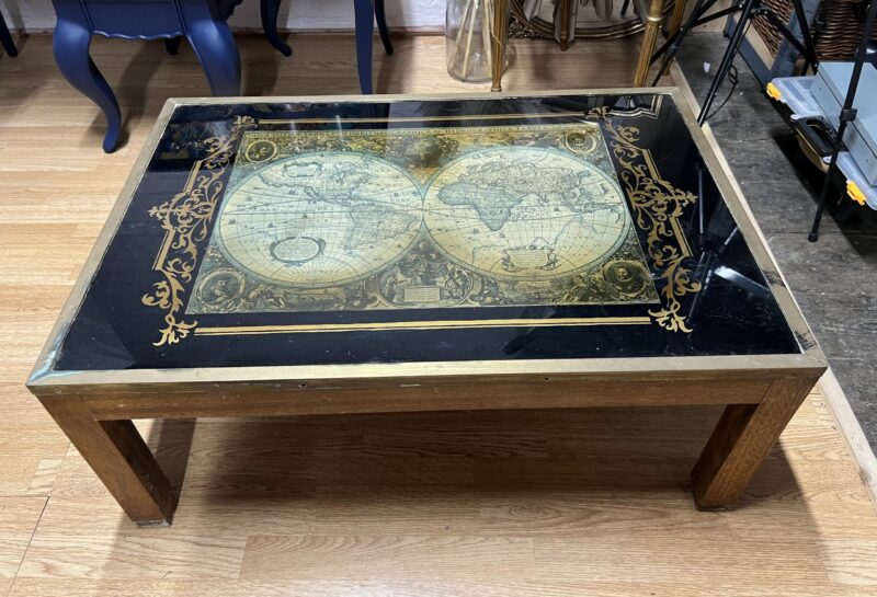 A wooden coffee table with a glass top featuring an intricate world map design. The map is bordered with ornate, golden patterns. The table is set on a polished wooden floor, with various pieces of furniture and objects visible in the background.