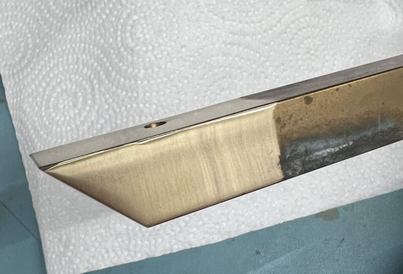 Image shows a metal blade with a sharp, polished edge placed on a white paper towel. The blade has a partially tarnished or oxidized section near the handle area. This setup is on a light blue surface.