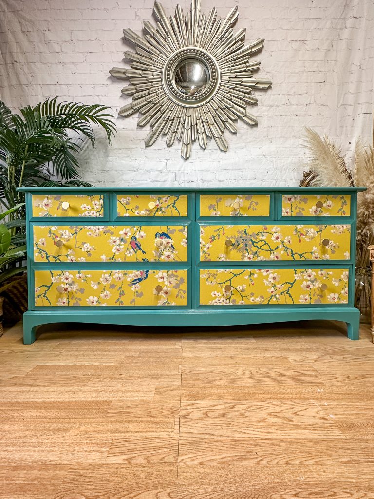 A wooden dresser with a teal frame and six yellow drawers featuring floral and bird illustrations. The dresser is set against a white brick wall, adorned with a decorative sunburst mirror. Potted plants and dried flowers are placed around the dresser.