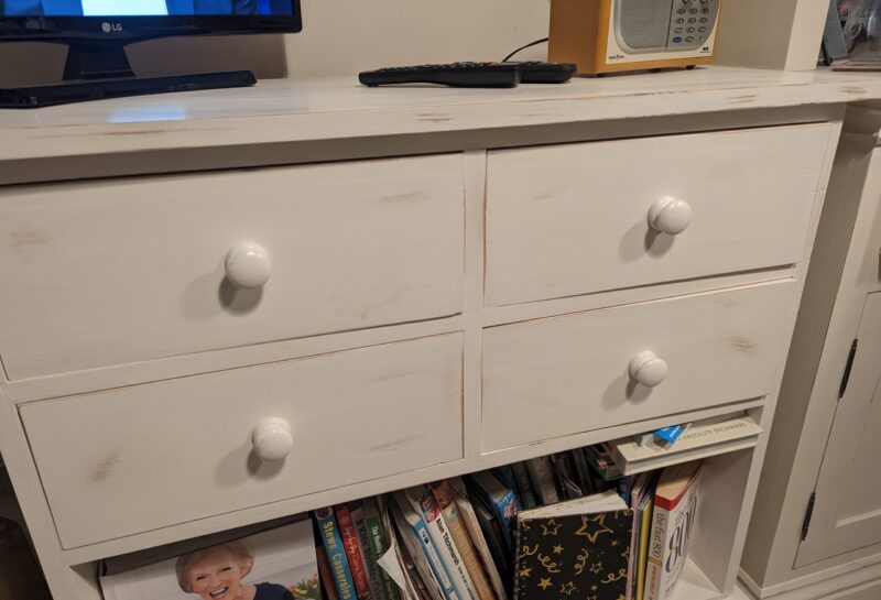 A white wooden dresser with four knobs and slightly distressed paint stands against a wall. On the left side of its bottom shelf, there's a pile of books, magazines, and a binder with a gold star pattern. A remote control sits on top beside a radio and a TV.