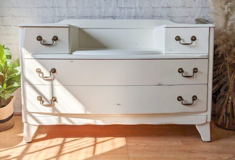 A white wooden dresser with three large drawers and two small compartments on top, each with brass handles. The dresser stands on a wooden floor in a room with white brick walls, a painting above, and some plants placed around it. Sunlight casts shadows on the floor.