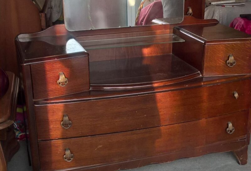 A vintage wooden dresser featuring a large rectangular mirror. The dresser has three drawers with metal handles and two smaller compartments on either side of the mirror. It is placed in a well-lit room with other furniture and decor visible in the background.