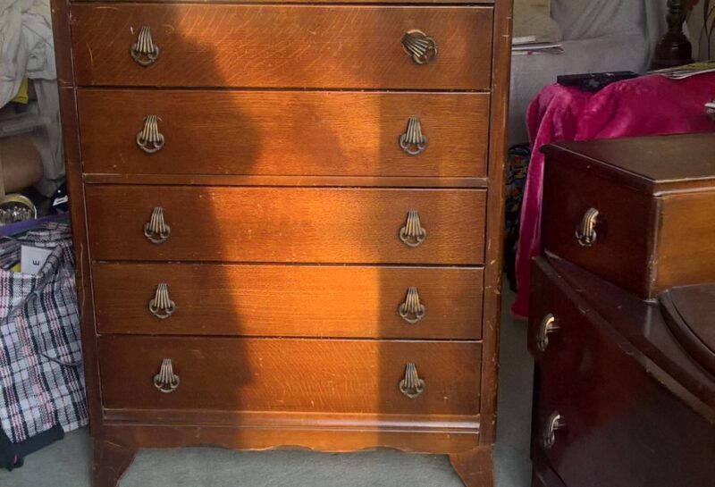 A wooden chest of drawers with brass handles stands in a living room. The chest has five drawers and is lit by sunlight from the left. Surrounding the chest are various items, including a red throw blanket on a nearby piece of furniture, a table lamp, and a painting on the wall.