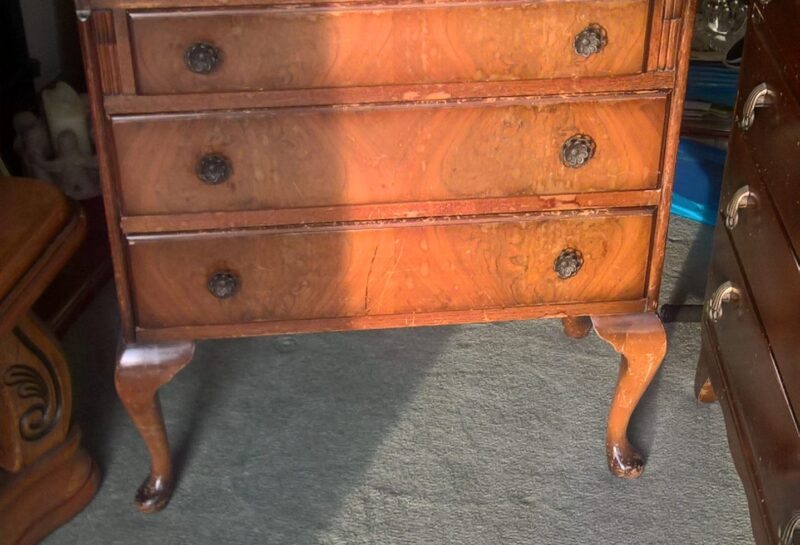 A wooden antique secretary desk with a fold-down writing surface and three drawers, standing on cabriole legs. The polished wood has a rich, warm finish, and decorative handles adorn each drawer. The desk sits on a gray carpeted floor in a room lit by sunlight.