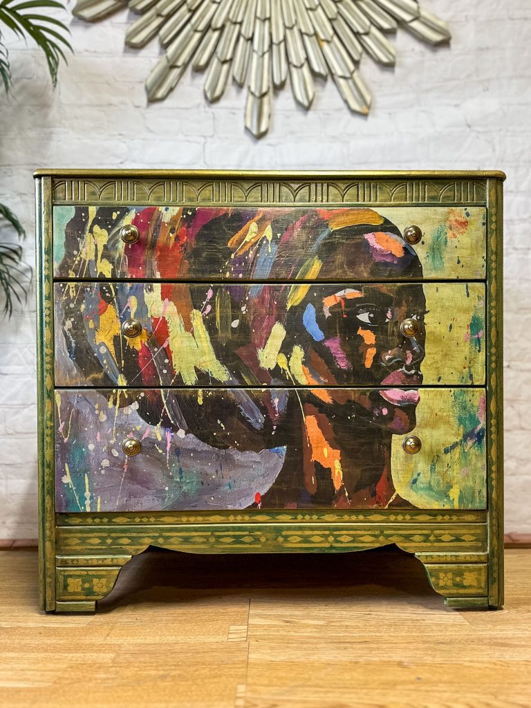 A painted dresser stands against a brick wall. The Bohemian Green & Gold: Revamped Harris Lebus MCM Chest of Drawers features a vibrant, abstract portrait of a person's profile on its front, highlighted with various colorful brush strokes. The green and gold frame with ornate details contrasts beautifully with the modern artwork. A decorative metal sunburst hangs above.