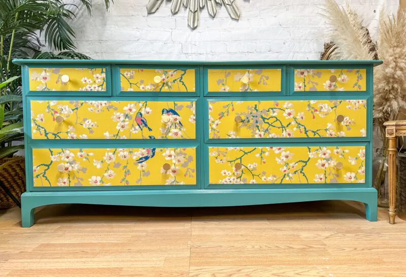 A turquoise dresser features nine drawers, each adorned with a vibrant yellow front. The yellow fronts are decorated with an intricate floral and bird pattern, adding a pop of color against the wooden floor. The background includes a white brick wall and green plants.