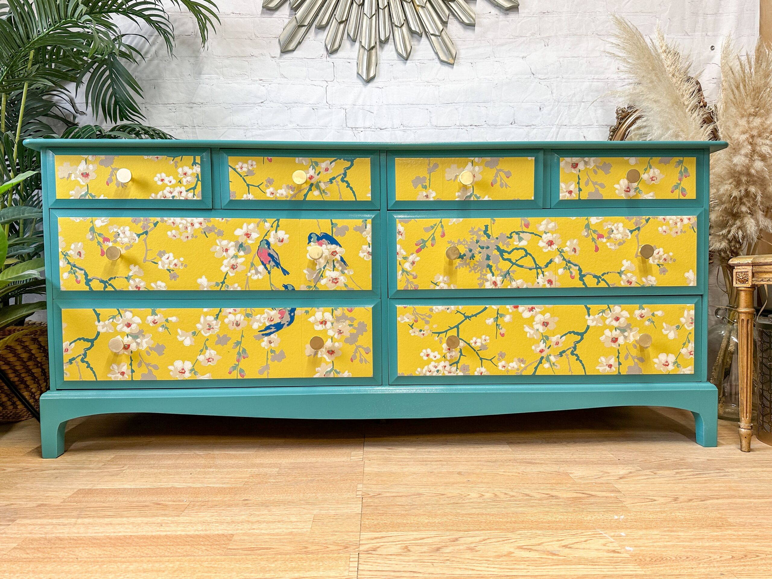 A turquoise dresser features nine drawers, each adorned with a vibrant yellow front. The yellow fronts are decorated with an intricate floral and bird pattern, adding a pop of color against the wooden floor. The background includes a white brick wall and green plants.