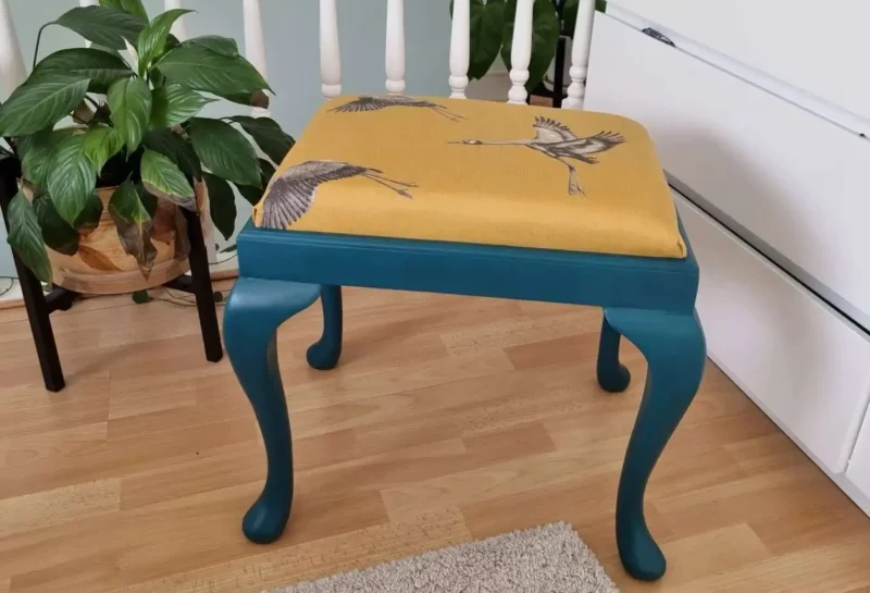 A blue wooden stool with a yellow cushion featuring bird illustrations is placed on a wooden floor. To its left is a potted plant with broad green leaves and in the background are white vertical railings and a white drawer unit. A beige rug is partially visible.