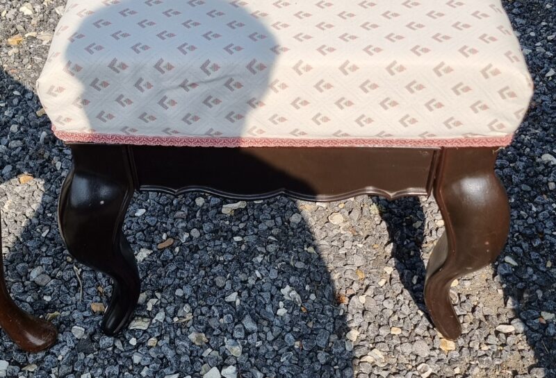 A small vintage stool with dark wooden, curved legs and a light-colored cushion featuring a repetitive pattern. The stool sits on a gravel surface outdoors, casting a shadow along with another shadow from a person taking the photograph.