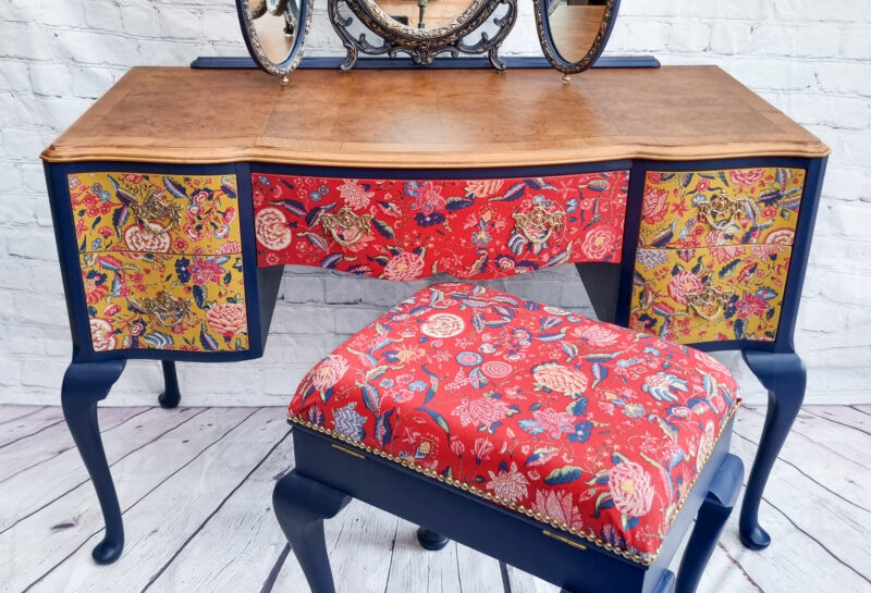 A vintage vanity set with an ornate triple mirror and colorful floral fabric on the drawers and matching stool. The vanity has dark blue legs and trim with a wooden tabletop. The background features a white brick wall and a wooden floor.