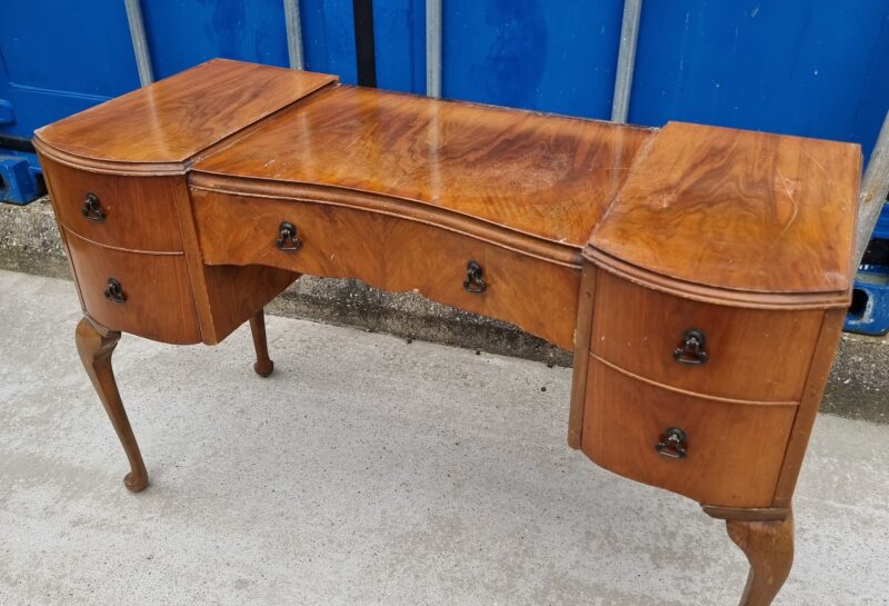 A vintage wooden desk with three drawers on each side. The desk features a curving design and ornate handles. It has cabriole legs and a polished surface, set against a backdrop of closed blue industrial containers.