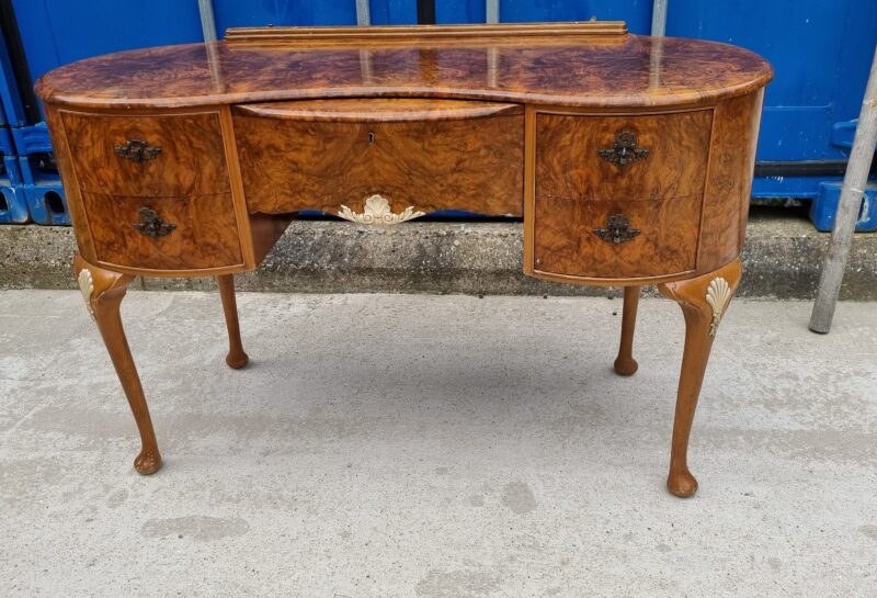 A vintage wooden dressing table with ornate carvings and a curved front. It has four small drawers with decorative handles and slender, cabriole legs. The table is placed on a concrete surface in front of a blue industrial background.