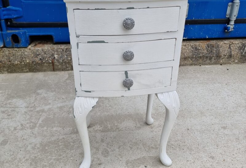 A small, white, vintage wooden nightstand with three drawers, each featuring a decorative knob. The piece has a slightly distressed look and stands on four elegantly curved legs. It's set against a background of a blue container and a concrete surface.