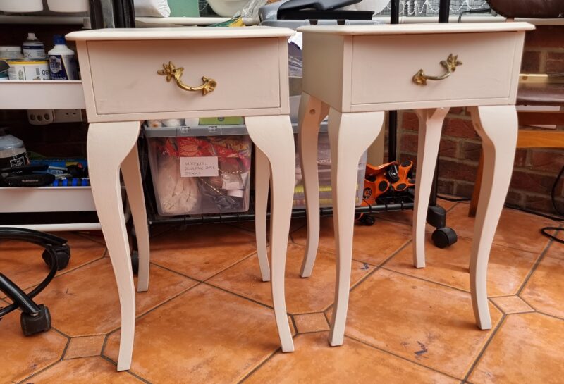 Two white, vintage-style side tables with single drawers stand on a tiled floor. Each table has cabriole legs and brass handles. The background includes various art supplies and equipment in a creatively cluttered workspace.