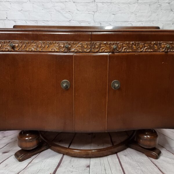 A vintage wooden cabinet with decorative carvings on the top edge and two front doors with circular handles. The cabinet has a polished finish and sits on a curved wooden base. The background features a white brick wall and a wooden floor.