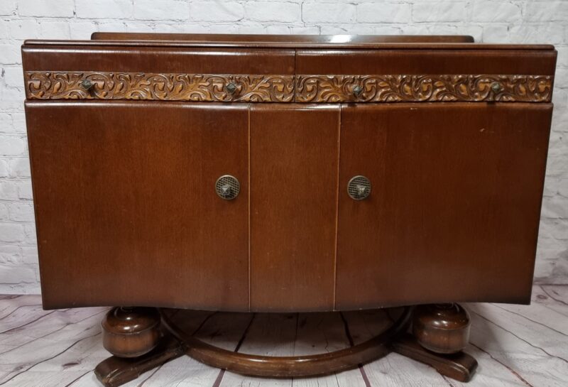 A vintage wooden cabinet with decorative carvings on the top edge and two front doors with circular handles. The cabinet has a polished finish and sits on a curved wooden base. The background features a white brick wall and a wooden floor.