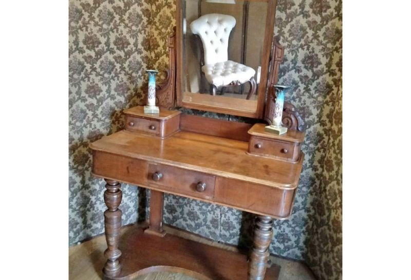 An antique wooden vanity with detailed carvings stands against a patterned wallpaper backdrop. The vanity features a large mirror, two small drawers on its surface, and two decorative vases. Reflected in the mirror is a white tufted chair.