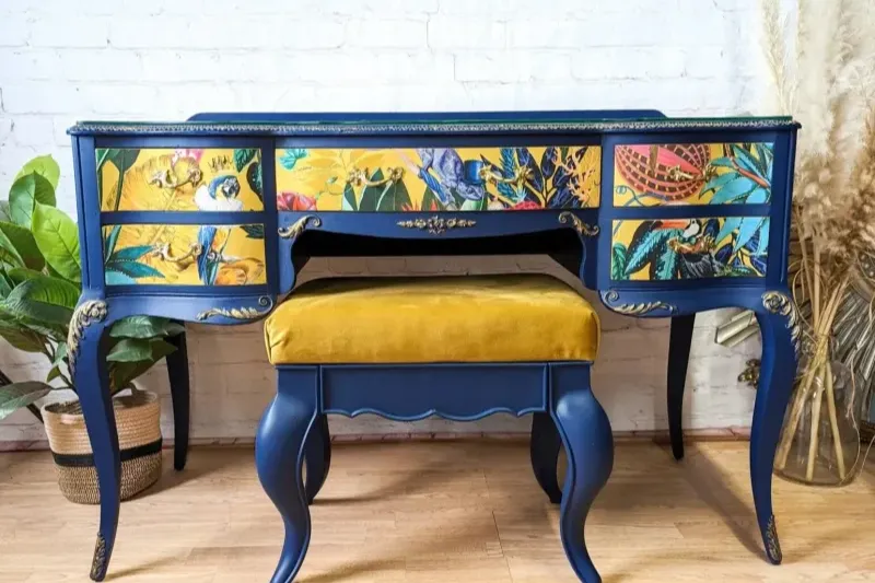 A vibrant blue vanity desk with an intricate floral and bird design on the drawers, paired with a matching blue stool that has a mustard-yellow cushion. The set is placed against a white brick wall with decorative dried plants and a potted plant nearby.