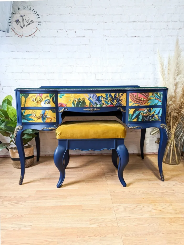 A blue vintage vanity with floral and bird designs on the drawer fronts. It features gold accents and ornate detailing. Paired with a mustard yellow upholstered stool, the set is placed against a white brick wall with dried pampas grass on the right side.