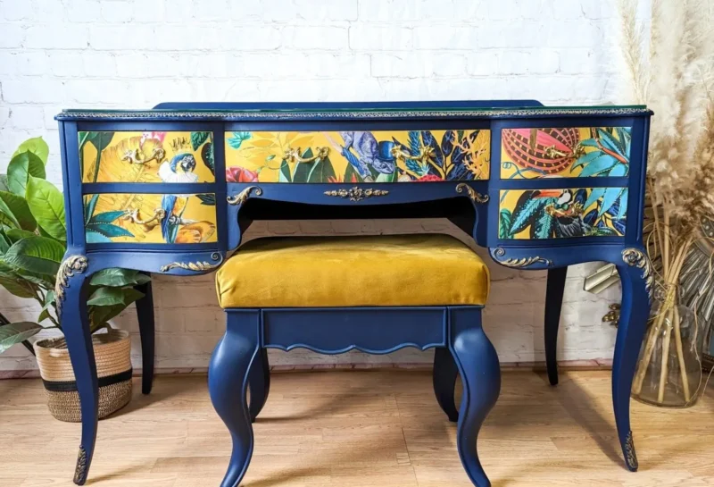 A blue vintage vanity with floral and bird designs on the drawer fronts. It features gold accents and ornate detailing. Paired with a mustard yellow upholstered stool, the set is placed against a white brick wall with dried pampas grass on the right side.