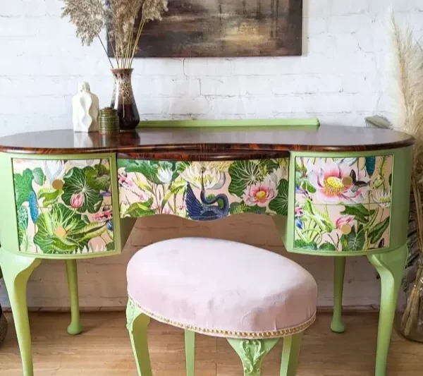 A vintage vanity desk with vivid, floral and bird-themed painted drawers sits against a white brick wall. The desk is light green with a wooden top and is accompanied by a cushioned, pale pink stool. Plants and decorative items surround the setup, adding to the aesthetic.