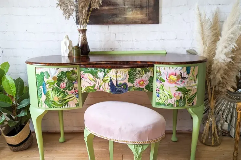 A vintage vanity desk with vivid, floral and bird-themed painted drawers sits against a white brick wall. The desk is light green with a wooden top and is accompanied by a cushioned, pale pink stool. Plants and decorative items surround the setup, adding to the aesthetic.