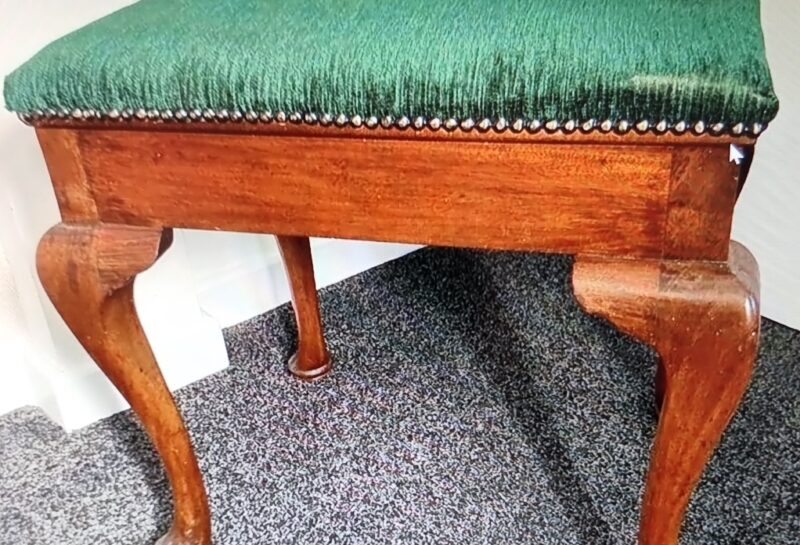 A wooden stool with ornate, carved legs and a green fabric cushioned seat. The edges of the seat are adorned with a row of metallic studs. The stool is positioned on a carpeted floor against a white and beige wall.