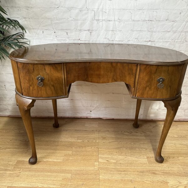 An antique wooden writing desk with curved edges, slender legs, and intricate metal handles stands on a hardwood floor. The background features a white brick wall, a decorative mirror, and green potted plants, creating a classic and elegant setting.