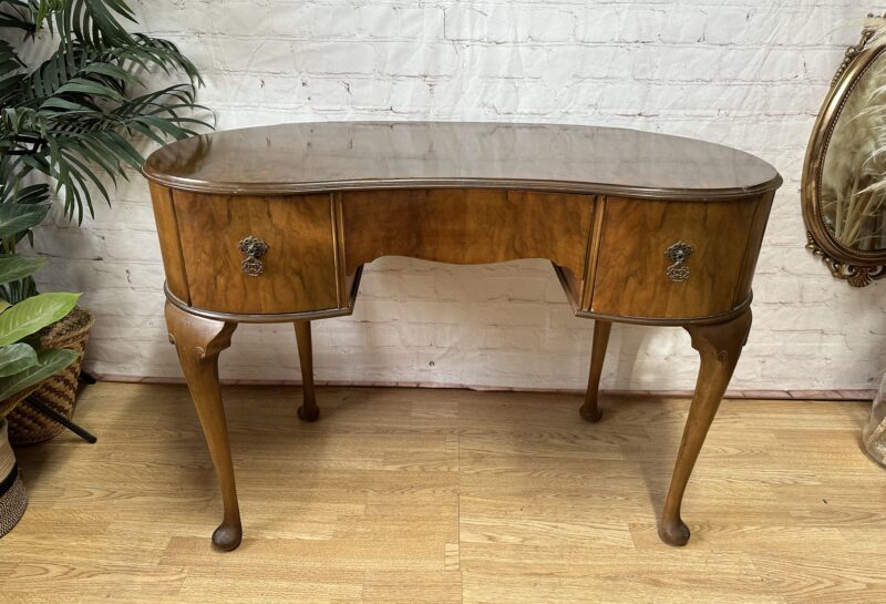 An antique wooden writing desk with curved edges, slender legs, and intricate metal handles stands on a hardwood floor. The background features a white brick wall, a decorative mirror, and green potted plants, creating a classic and elegant setting.