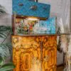 A vintage wooden cabinet with ornate grain patterns and decorative handles is topped with an open bar compartment displaying a variety of glassware and bottles. The interior is lined with blue wallpaper featuring a geometric design. Plants and a framed picture are nearby.