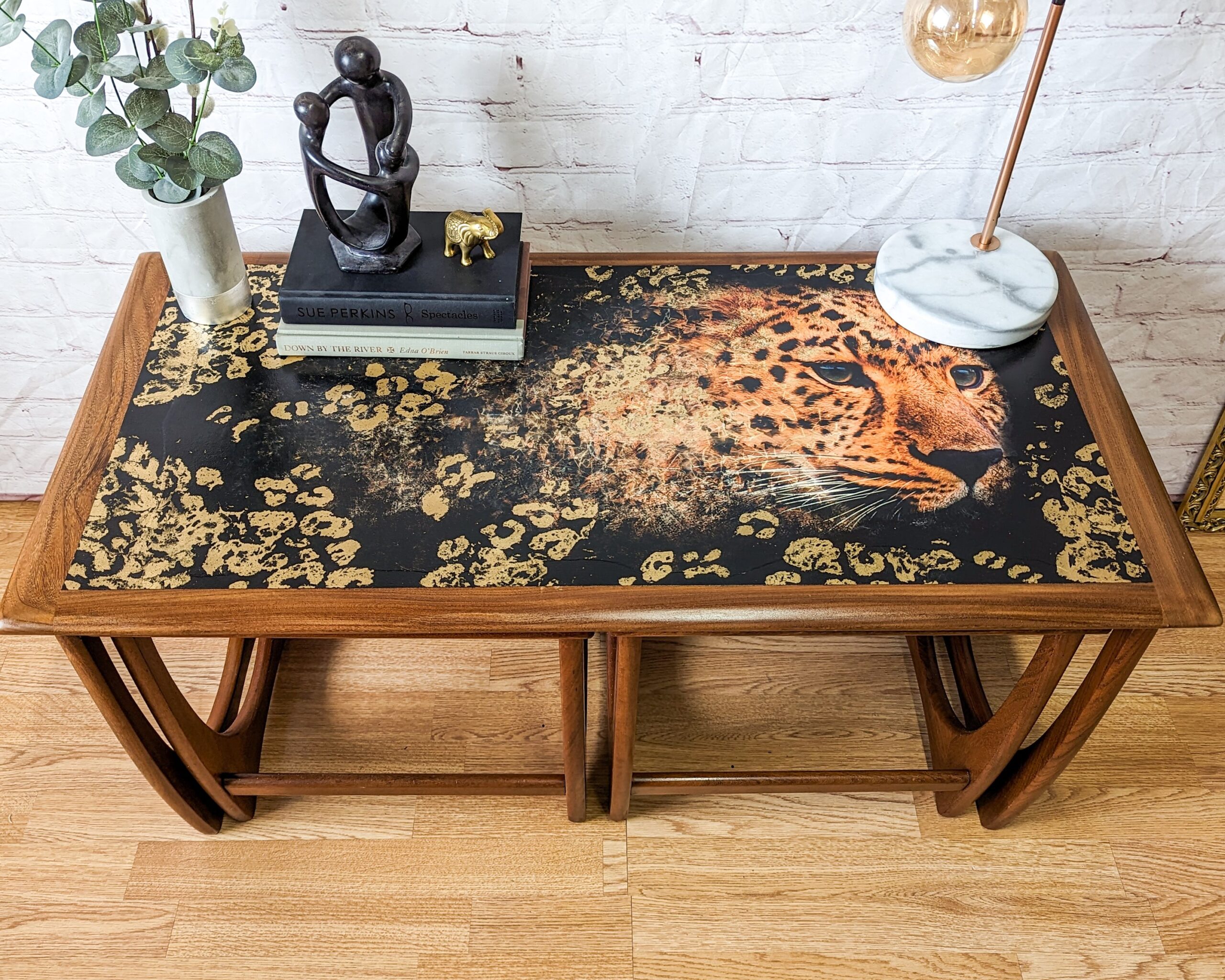 A wooden table with a painted surface featuring a detailed image of a leopard's head surrounded by fading spots. On the table are decorative items including a modern sculpture, small gold figurine, books, a plant, and a lamp with a marble base.