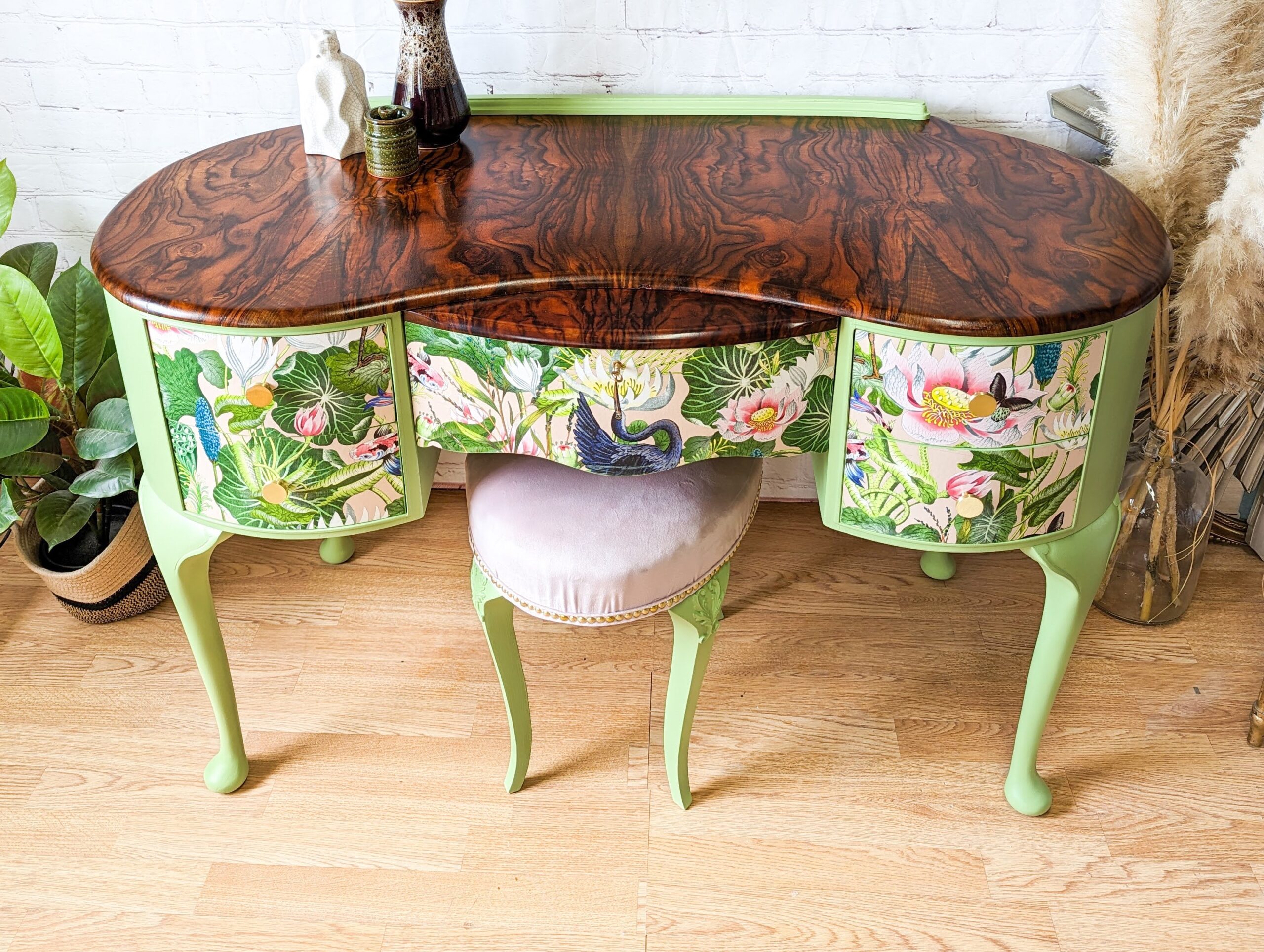 An elegant vintage vanity with a wooden top and pastel green legs, adorned with nature-themed decals featuring birds and foliage. A plush, light pink cushioned stool sits underneath. Potted plants and decorative items are arranged in the background.