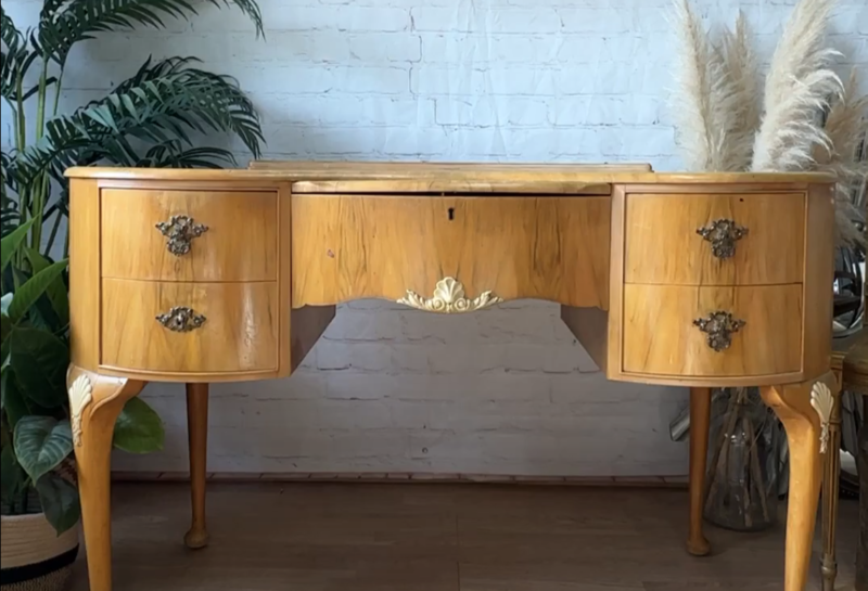 A vintage wooden dressing table with ornate legs and handles, featuring four curved drawers. In the background, a white brick wall adorned with a framed painting of a bull, a potted plant on the left, and decorative pampas grass on the right.