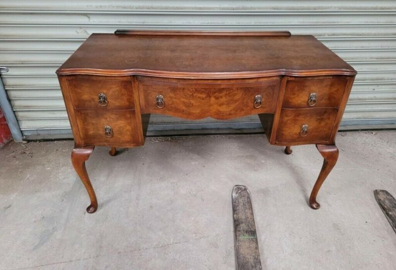 A vintage wooden desk with an ornate design stands in front of a metal garage door. The desk features curved legs, two drawers on each side, and a central drawer. It has brass handles and a darker wood inlay on the front. The desk shows signs of wear.