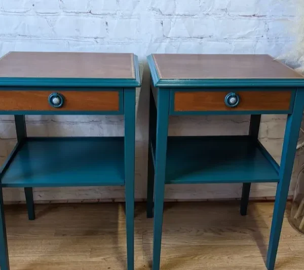 Two matching teal-painted wooden side tables with single drawers featuring brown fronts and round handles. Each table has a lower shelf for storage. They are positioned side by side on a wooden floor, against a white brick wall, with plants and dried pampas grass nearby.