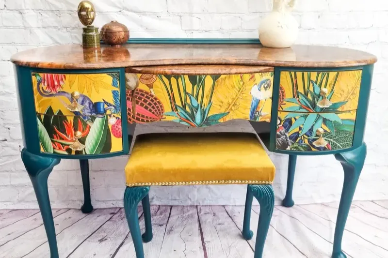 A vibrant desk features a colorful tropical-themed design on the front panels, depicting birds and plants. The desk has teal legs and a wooden top. In front of the desk is a mustard yellow cushioned stool with studded trim. Decorative items are placed on top of the desk.