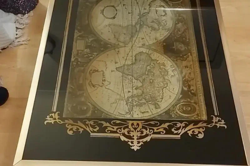 A framed antique-style world map with two hemispheres in sepia tones, bordered by an ornate gold design. The map appears to be encased in glass, reflecting light. The frame is a simple, elegant gold. The background surface is light wood.