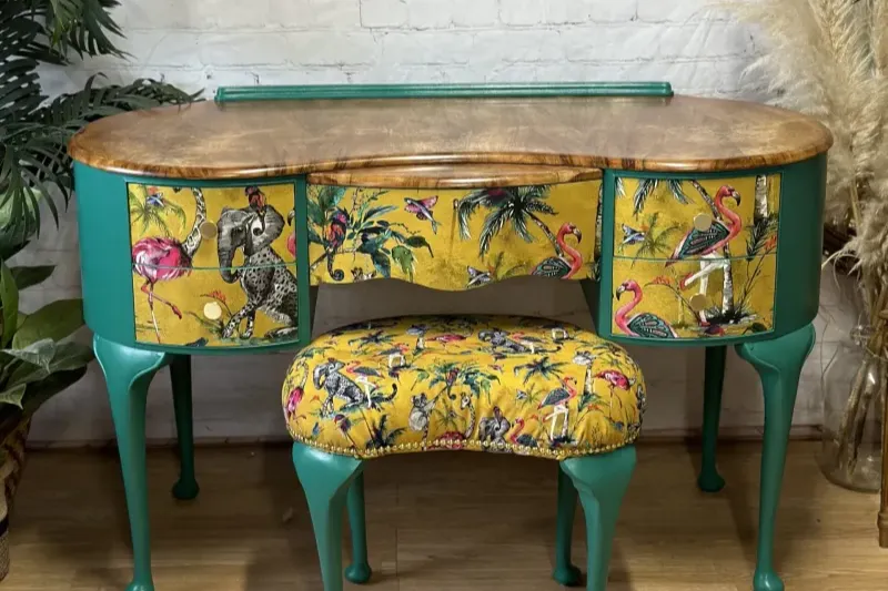 A vibrant wooden vanity with a matching stool, both painted teal. The vanity and stool are adorned with colorful bird-themed fabric featuring flamingos, parrots, and cranes, set against a yellow background. The setup is placed on a wooden floor with green plants nearby.