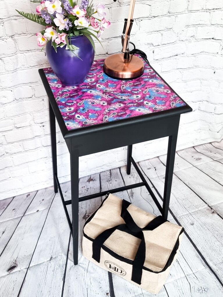 A small table with a colorful, intricate patterned top, holding a purple vase filled with flowers and a copper lamp. Underneath the table is a beige and black insulated bag. The setup is against a white brick wall and wooden floor.