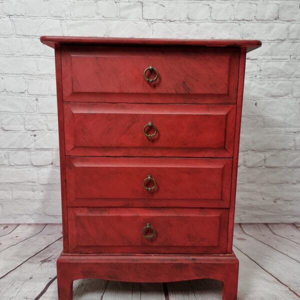 A small red wooden dresser with four drawers and brass ring handles stands on a wooden floor against a white brick wall background.