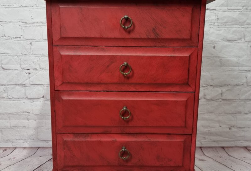 A small red wooden dresser with four drawers and brass ring handles stands on a wooden floor against a white brick wall background.